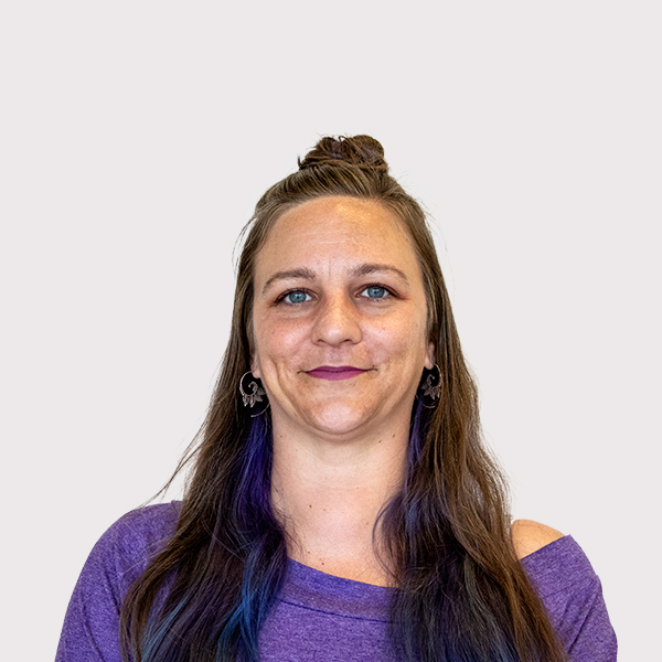 A woman with long hair wearing a purple top and earrings, smiling against a neutral background.